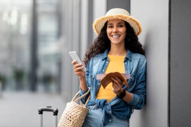 Smiling attractive brunette young woman tourist holding flight tickets, passport and smartphone, checking her flight information on Internet, standing by airport with suitcase, copy space