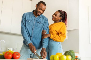 Loving young black man cooking delicious dinner for his beautiful wife, cutting vegetables, light kitchen interior, copy space. Love, affectionate, family lifestyle concept