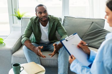 Black man suffering from depression, counselor providing professional assistance at medical office. Young male patient having consultation with psychologist, receiving help