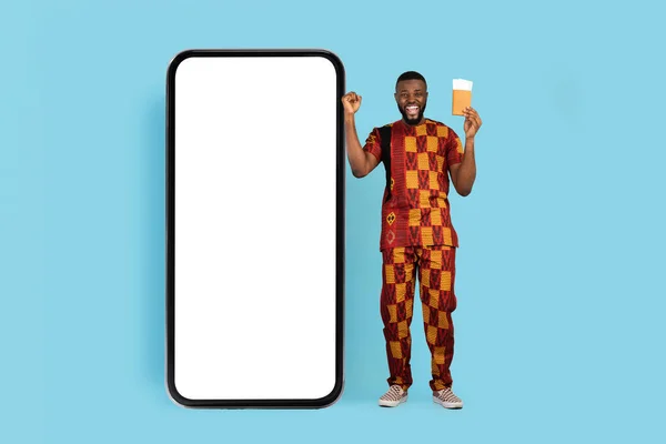 Viza Center Ad. Excited Black Man With Pasport And Tickets Standing Near Huge Blank Smartphone Over Blue Background, African Guy In Traditional Costume Recommending Online Services, Mockup