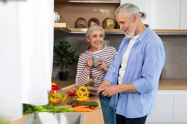 Happy Senior Couple Relaxing At Home, Cooking Healthy Food And Drinking Tea In Kitchen Interior, Cheerful Elderly Couple Preparing Fresh Vegetable Salad, Man Chopping Bell Pepper