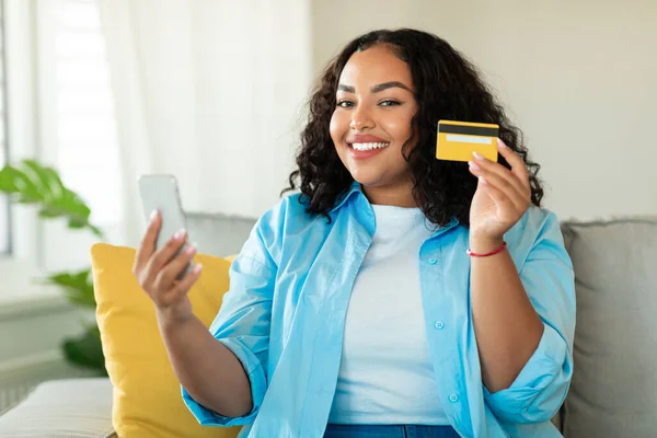 Mobile Shopping. Cheerful Black Woman Using Cellphone And Credit Card Making Payment Purchasing Plus Size Clothes Sitting On Couch At Home, Smiling To Camera. Ecommerce Concept. Selective Focus
