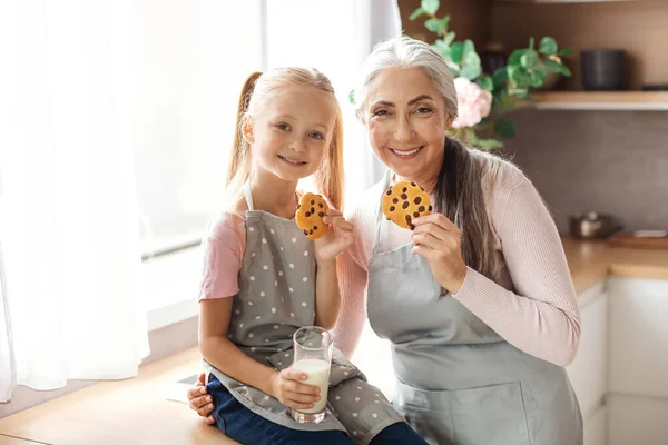 Smiling European Granddaughter Old Grandma Aprons Enjoy Cookies Chocolate Glass — Foto de Stock