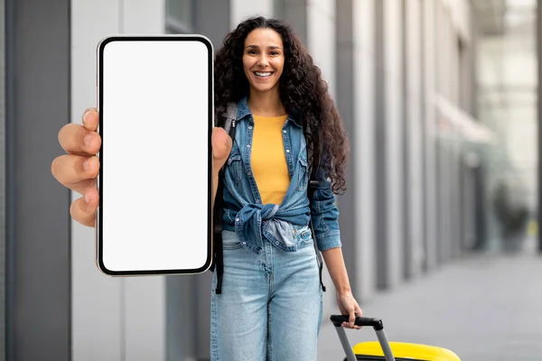 Cheerful pretty millennial woman in casual tourist walking by airport with luggage, showing cell phone with white empty screen, recommending nice online travelling offer, mockup, copy space