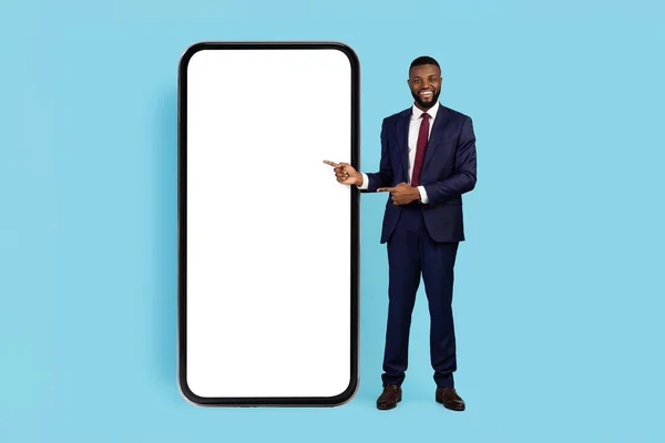 Check This. Handsome Black Businessman Wearing Suit Pointing At Big Blank Smartphone With White Screen, African American Male Entrepreneur Showing Copy Space While Posing On Blue Background, Mockup