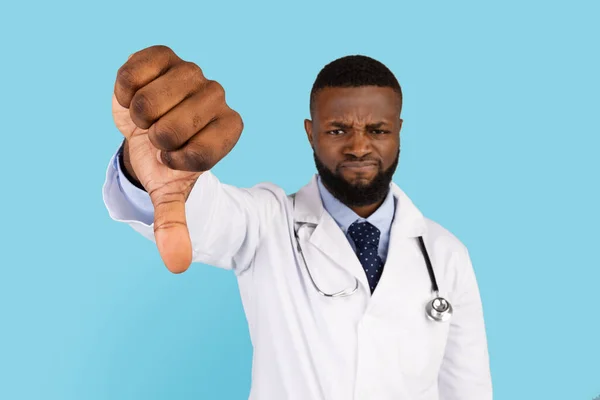 Displeased Black Male Doctor Showing Thumb Down Gesture At Camera, Upset Young African American Medical Worker In White Coat Expressing Negotive Emotion, Standing Isolated On Blue Background