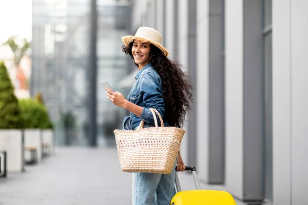 Smiling young lady tourist in casual outfit and straw hat going to airport, carrying big yellow suitcase, holding summer bag and smartphone, check-in online, panorama with copy space