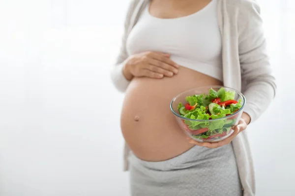 Nutrition and healthy diet during pregnancy concept. Unrecognizable pregnant woman holding bowl with fresh vegetable salad and touching her big tummy, white background, cropped, free space