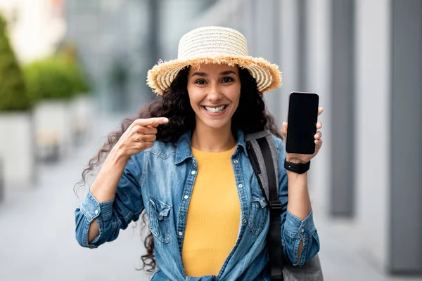 Cheerful pretty millennial woman in casual tourist walking by airport, pointing at smartphone with black empty screen in her hand, recommending nice online travelling offer, mockup, copy space