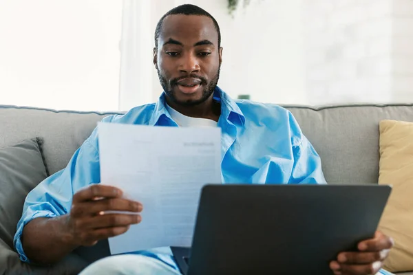 Remote career, freelance job concept. Young african american man using laptop computer and looking through business documents during online work at home