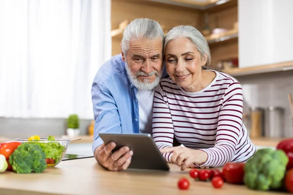 Portrait Of Happy Elderly Spouses Using Digital Tablet In Kitchen Interior, Smiling Married Senior Couple Relaxing With Modern Gadget, Checking Food Recipe Or Reading News Online, Copy Space