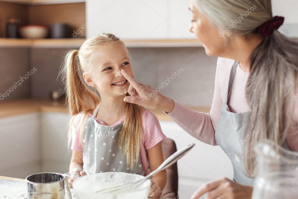 Happy senior woman in apron touches nose of little girl, make dough and have fun with flour, family enjoy leisure time in kitchen interior. Cooking lesson baking and prepare homemade food together