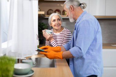 Caring Elderly Husband Helping Wife With Dishes In Kitchen, Smiling Senior Man Cleaning Plates While His Spouse Drinking Coffee, Happy Aged Spouses Sharing Domestic Chores, Free Space
