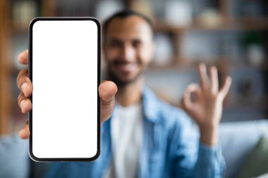 Mobile Offer. Smiling black man demonstrating big smartphone with blank white screen and showing ok gesture at camera while sitting on couch at home, recommending new app or website, collage, mockup