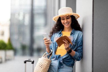 Happy beautiful young woman in casual outfit and straw hat standing by airport with luggage and summer bag, check-in online, using cell phone and looking at flight ticket, copy space
