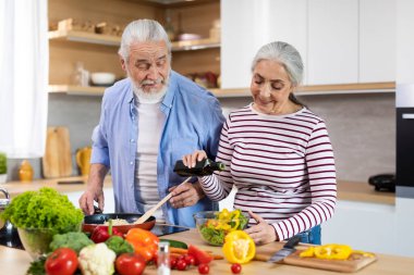 Happy Elderly Couple Preparing Tasty Lunch Together In Kitchen, Smiling Senior Man And Woman Cooking Healthy Vegetarian Meal At Home, Aged Woman Adding Oil To Fresh Vegetable Salad, Free Space