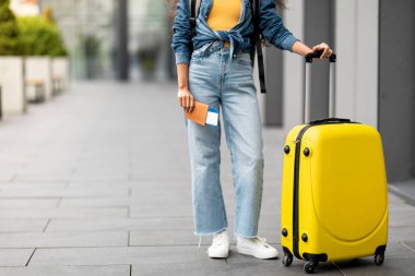 Cropped of woman in casual outfit tourist with yellow suitcase luggage holding passport and tickets, posing in airport, panorama with copy space. Travelling, tourism, vacation, holidays concept