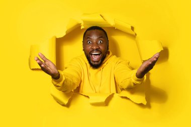 Excited african american man spreading hands sideways, looking at camera with open mouth through yellow paper hole, banner, copy space