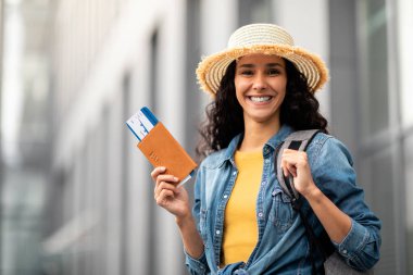 Closeup of cheerful pretty long-haired brunette millennial woman holding passport and flight tickets, smiling at camera, carrying backpack, wearing summer hat, going vacation, copy space