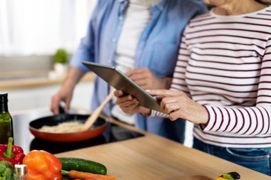 Senior Couple Using Digital Tablet In Kitchen While Cooking Food, Unrecognizable Elderly Man And Woman Browsing Internet On Modern Gadget While Preparing Tasty Meal At Home, Closeup, Cropped Shot