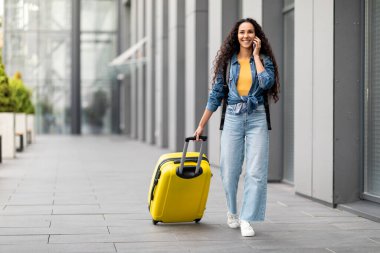 Smiling young brunette curly woman in casual outfit traveller walking by airport, carrying yellow suitcase, having phone conversation, looking at copy space, panorama, full length