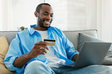 E-commerce. Happy black man holding credit card and shopping online with laptop computer, sitting on couch at home. Male customer showing bankcard, copy space