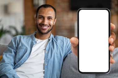 Place For Ad. Cheerful Black Man Demonstrating Blank Smartphone With Big White Screen At Camera While Sitting On Couch At Home, African American Male Recommending New Mobile App, Collage, Mockup