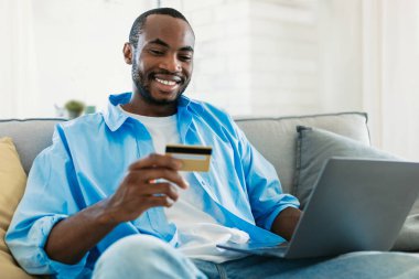 Black male bank client looking at plastic credit card in hand and entering information using pc laptop, shopping online in webstore, purchasing goods or services