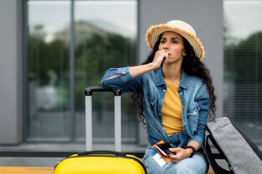 Commom travel problems concept. Pensive pretty long-haired brunette young woman in straw hat tourist sitting on bench with backpack and suitcase outdoors, holding passport and cell phone, copy space
