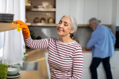 Portrait Of Smiling Senior Woman Cleaning Shelves In Kitchen From Dust, Tidy Elderly Female With Rag In Hand Polishing Surface, Enjoying Doing Domestic Chores And Cleaning Home, Closeup
