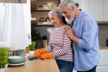 Portrait Of Happy Senior Spouses Washing Dishes In Kitchen After Lunch, Loving Elderly Husband Embracing Wife From The Back While She Doing Domestic Chores, Side View With Free Space