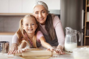 Happy retired woman in apron teaching little girl doing dough using rolling pin making cookies in minimalistic kitchen interior. Household chores together, prepare food and cooking lesson at home