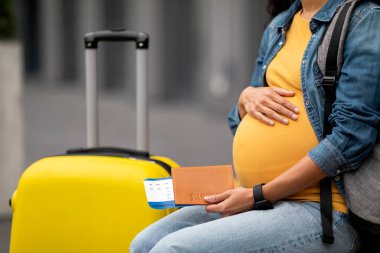 Birth tourism concept. Unrecognizable expecting woman travelling alone, sitting on bench with backpack next to yellow suitcase, holding passport, flight tickets, hugging her big belly, cropped