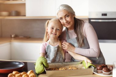 Cheerful european little granddaughter and elderly grandmother in aprons and mittens show baking sheet with cookies with chocolate on kitchen interior. Cooking baking for family at home, homemade food