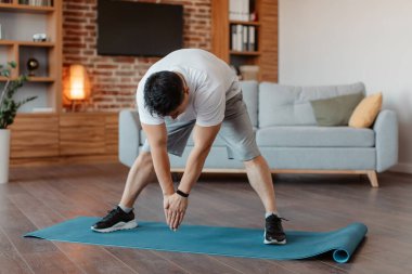 Active asian mature man doing exercises and stretching legs muscles at home, practicing domestic sports, full length. Middle aged man exercising in living room interior