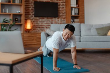 Online fitness class concept. Athletic asian man planking on yoga mat at home, watching fitness video on laptop, living room interior, copy space