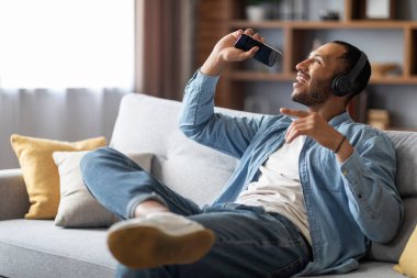 Domestic Fun. Cheerful Black Guy Listening Music In Wireless Headphones And Singing At Home, Positive Young African American Man Sitting On Couch With Smartphone, Enjoying Favorite Playlist