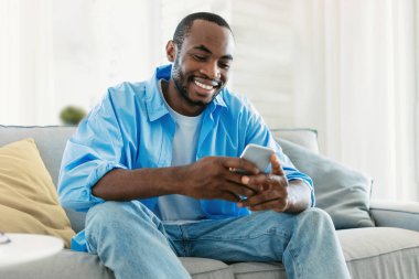 Excited black man chatting on smartphone, watching video, playing game while resting on sofa in living room interior. Free time for blog at home, new app, advertisement and offer
