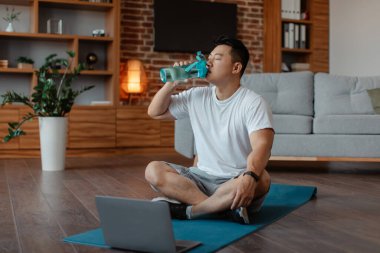 Sporty asian mature man sitting on mat with laptop, taking break from domestic workout, drinking water, copy space. Male keeping hydrated during online yoga training