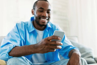 Happy african american man using smartphone, browsing social network, watching video or chatting with friends at home, resting on sofa, copy space, selective focus