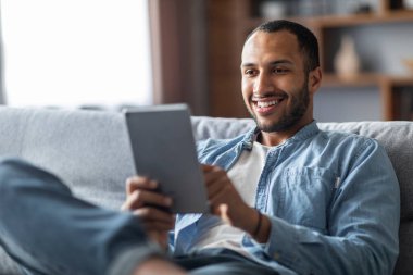 Handsome smiling black male relaxing with digital tablet at home, young african american man resting on couch and using modern gadget, browsing internet or shopping online, closeup shot