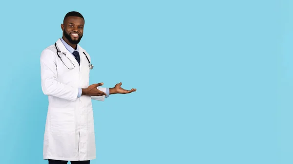Medical Ad. Smiling black male doctor pointing aside at copy space, young african american physician wearing uniform standing isolated over blue background, panorama with free place for design