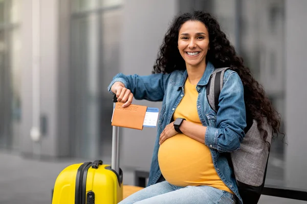 Giving birth in foreign country concept. Happy young long-haired brunette pregnant woman travelling alone, sitting on bench with backpack next to baggage, holding passport, flight tickets, copy space