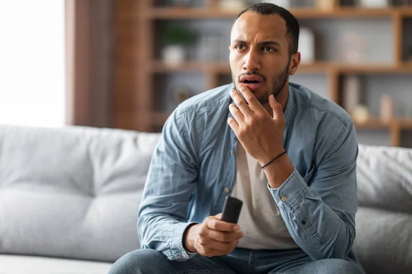 Shocked Black Man With Remote Controller In Hand Sitting On Couch At Home, Confused Young African American Guy Watching Tv Show In Living Room, Emotionally Reacting To Shocking Content, Copy Space