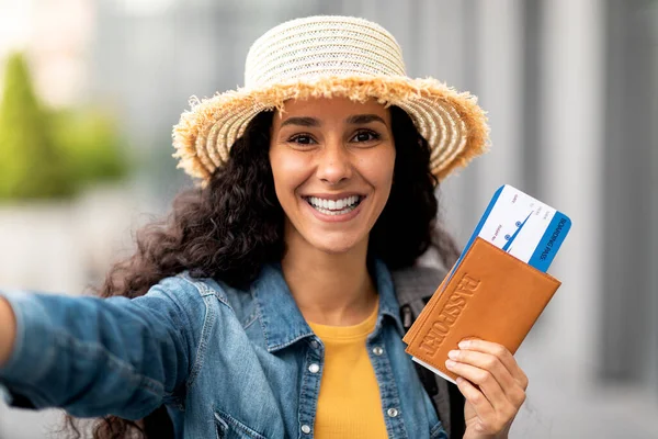 Excited beautiful middle eastern young woman in straw hat taking selfie, showing flight tickets and passport, sharing good news on social media, happy lady going summer vacation abroad, closeup