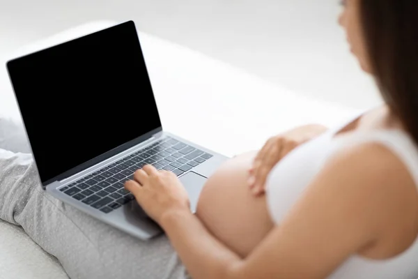 Unrecognizable pregnant lady using laptop with black empty screen, working in homeoffice, taking online education courses during maternity leave, touching her belly, closeup, mockup, bedroom interior