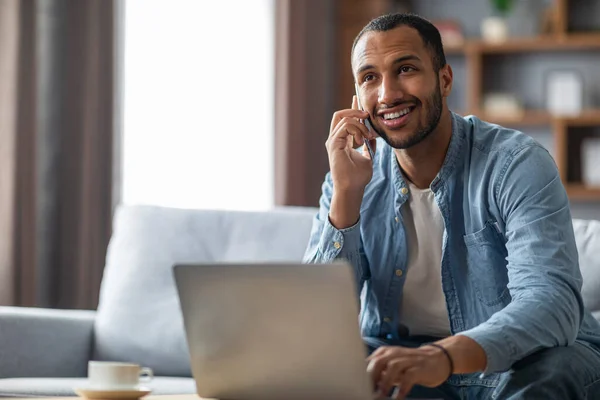 Handsome Young Black Man Talking On Cellphone And Using Laptop At Home, Smiling African American Man Sitting On Couch, Working Remotely With Computer Or Making Online Purchases, Copy Space