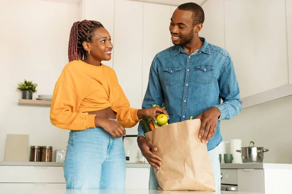 Ordering food online. Happy african american couple arriving from supermarket with grocery bag and unpacking in kitchen, looking at each other and smiling