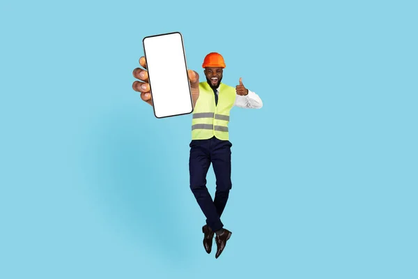 Cool Promo. Overjoyed Black Engineer With Blank Smartphone In Hand Jumping And Showing Thumb Up, Happy Excited African American Builder Demonstrating Free Space For Mobile Advertisement, Mockup