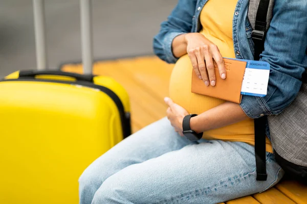 Giving birth abroad concept. Unrecognizable pregnant woman travelling alone, sitting on bench with backpack next to baggage, holding passport, flight tickets, hugging tummy, copy space, cropped